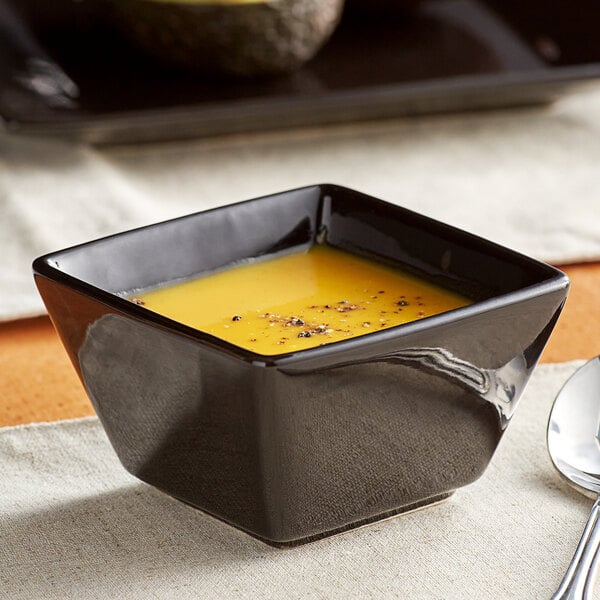 A bowl of soup in an Acopa Glossy Black Stoneware Bouillon Cup with a spoon on a table.