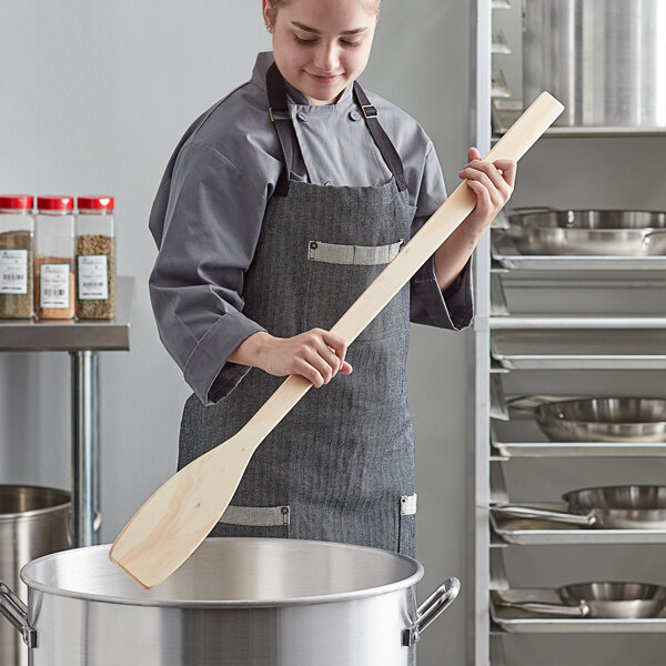 A woman in a chef's uniform using an American Metalcraft wood paddle to stir a pot of soup.