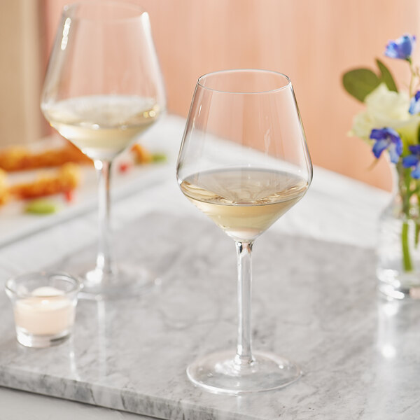 Two Acopa Silhouette wine glasses filled with white wine on a marble table with flowers.