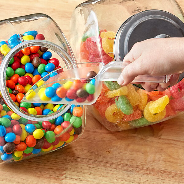 A hand using a Carnival King clear plastic utility scoop to pour yellow candy into a jar.
