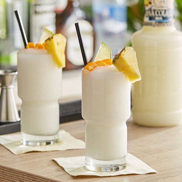 A close-up of a white bottle of Finest Call Premium Pina Colada Mix with two drinks garnished with pineapple and orange slices.