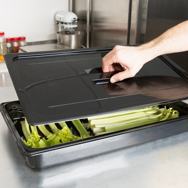 A hand using a black Cambro lid to open a tray of celery.