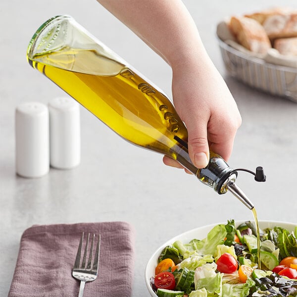 A hand pours olive oil from a Tablecraft green tinted glass oil cruet into a bowl of salad.