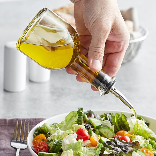 A hand pouring oil into a bowl of salad using a Tablecraft clear glass oil cruet.