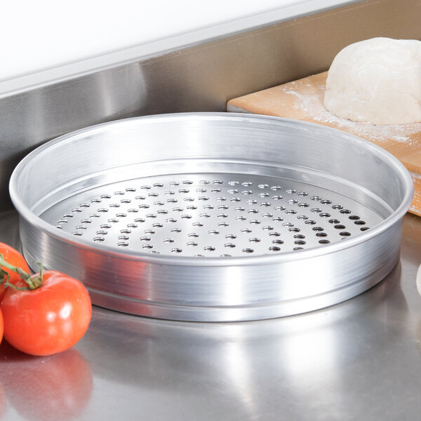 An American Metalcraft heavy weight aluminum pizza pan with holes next to a ball of dough and tomatoes.