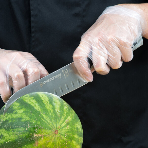 A person in gloves uses a Dexter-Russell V-Lo Santoku knife to cut a watermelon on a counter.