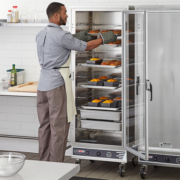 A man in a professional kitchen using a ServIt holding cabinet with a clear door to put trays of food inside.