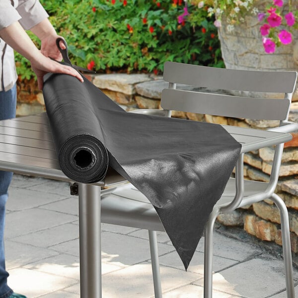 A person rolling a Choice black vinyl table cover roll on a table.