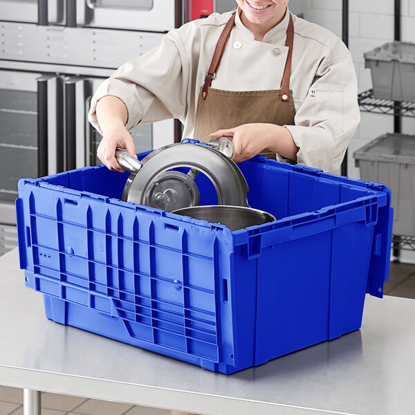 A woman in a chef's uniform putting a pan in a blue Orbis Stack-N-Nest tote with a metal lid.