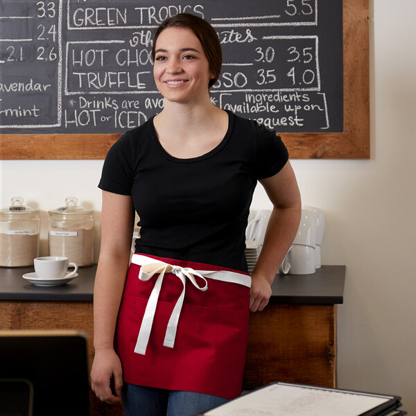 A woman wearing a Choice red poly-cotton waist apron standing in front of a chalkboard.