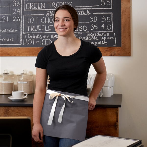 A woman wearing a gray Choice poly-cotton waist apron with natural webbing standing in front of a chalkboard.