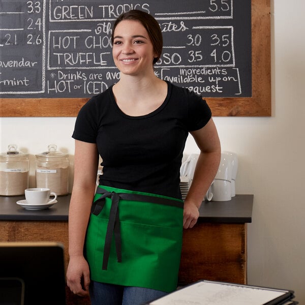 A woman wearing a Choice Kelly Green poly-cotton waist apron with black webbing standing in front of a chalkboard.