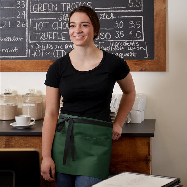 A woman wearing a black shirt and a hunter green waist apron with black webbing standing in front of a chalkboard.
