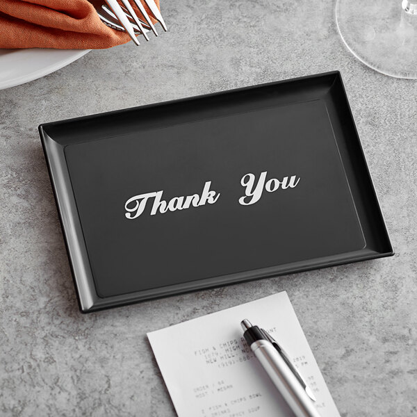 A black rectangular Vollrath Thank You Tip Tray with silver text on it next to a pen and paper.