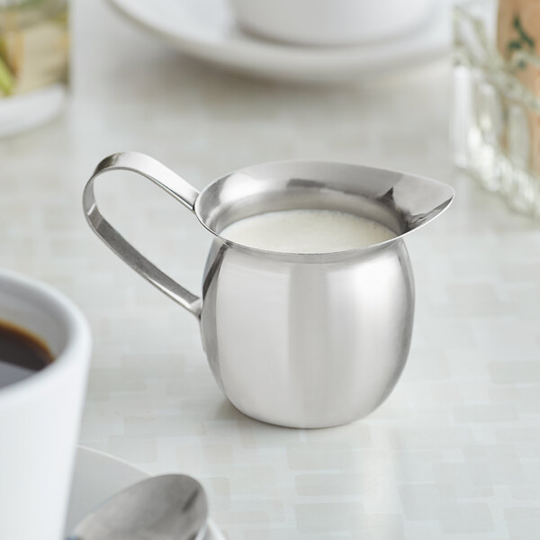 A Vollrath stainless steel bell creamer with a white liquid and a spoon.