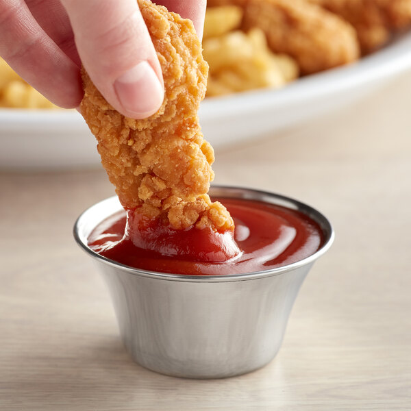 A hand holding a piece of fried chicken dipping into a Vollrath stainless steel sauce cup filled with ketchup.