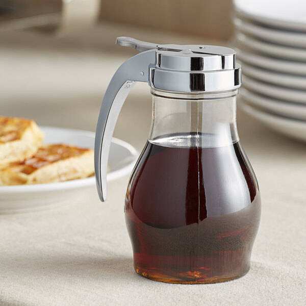 A Vollrath clear polycarbonate teardrop syrup server jar on a table of food with brown syrup inside.
