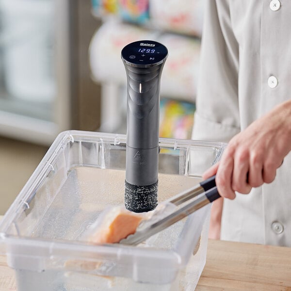 A person using a Galaxy SV100 sous vide immersion circulator to cook a piece of food in a container.