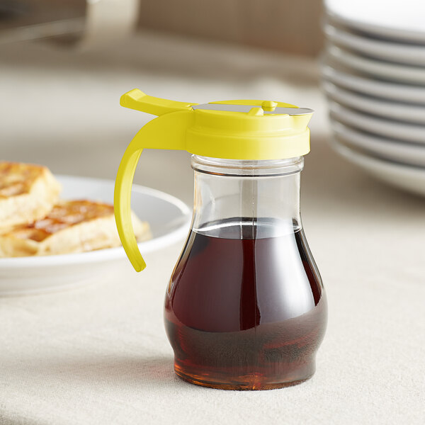A Vollrath clear polycarbonate teardrop syrup server with yellow top on a table with syrup in it.