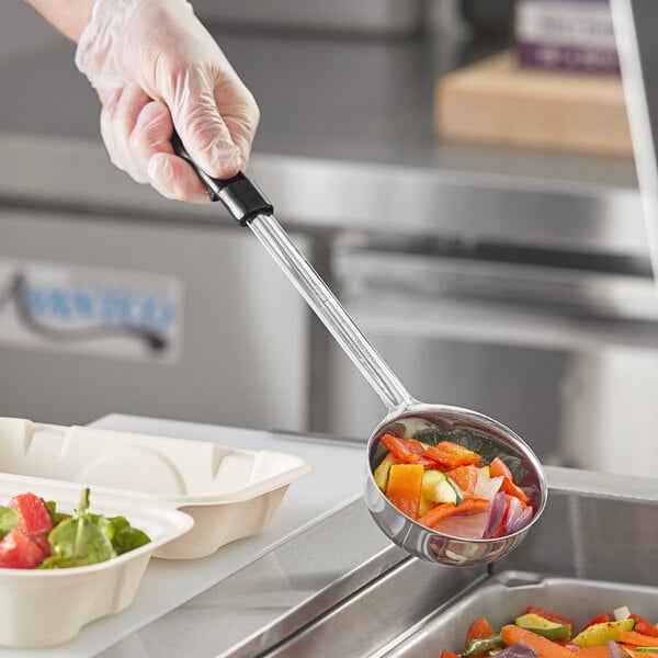 A hand in a plastic glove holding a black Choice portion spoon over a bowl of vegetables.
