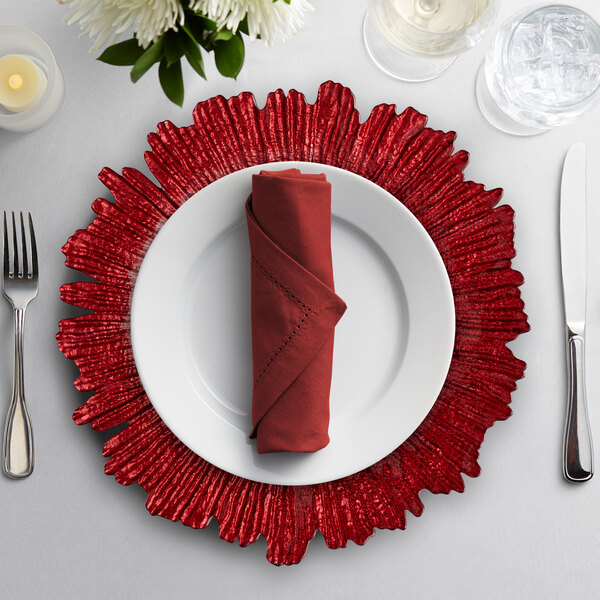 A red 10 Strawberry Street glass charger plate on a table with a napkin, fork, and knife.