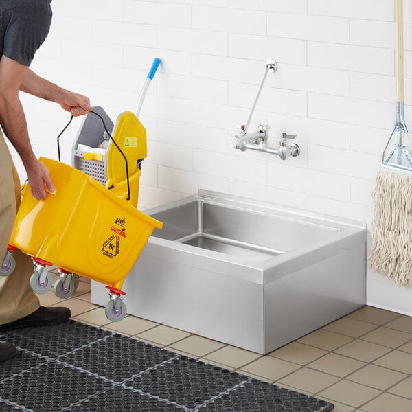 A man using a yellow mop to clean a Regency stainless steel floor mop sink.