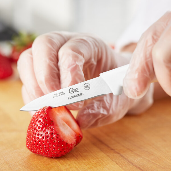 A person in gloves uses a Choice 3" Smooth Edge Paring Knife with a white handle to cut a strawberry.