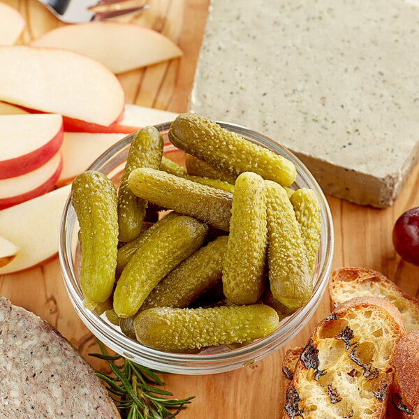 A bowl of pickles next to a block of cheese on a table with bread and fruit.