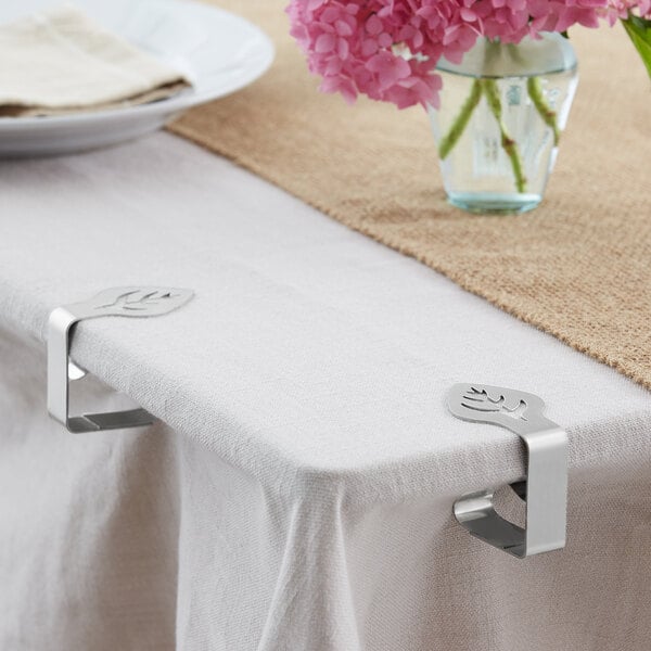 A table with a white tablecloth and a Choice stainless steel leaf design tablecloth clip with a vase of pink flowers.