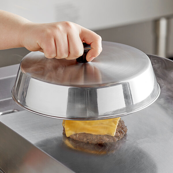 A hand holding a stainless steel basting cover over a hamburger.