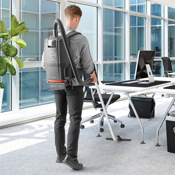 A man using a Hoover cordless backpack vacuum cleaner to clean an office.