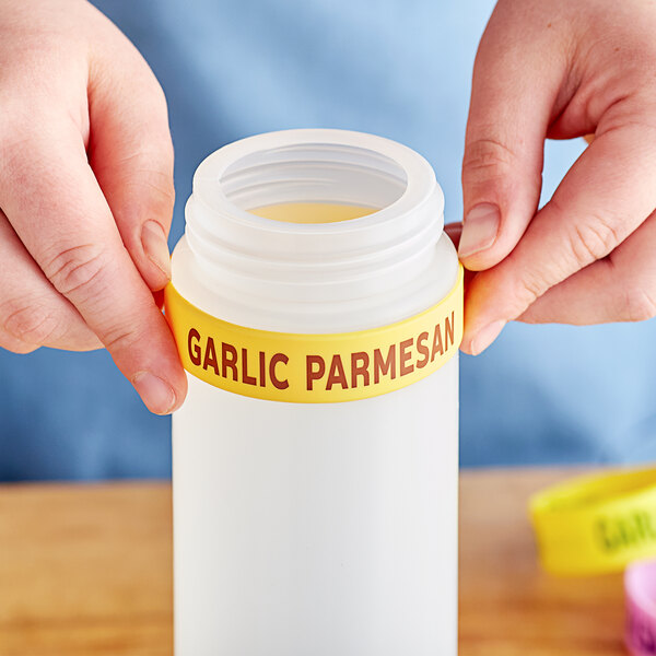 A person holding a white silicone label band with black text that says "Garlic Parmesan" over a yellow background on a jar.