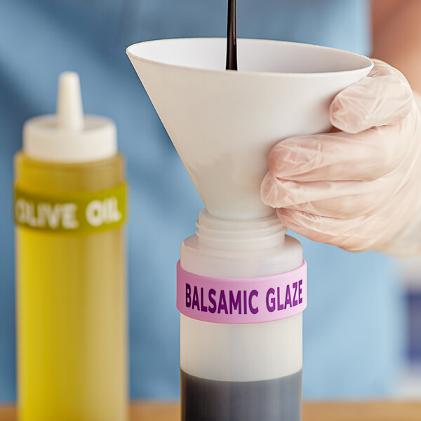 A hand using a funnel to pour balsamic glaze into a squeeze bottle with a white label band.