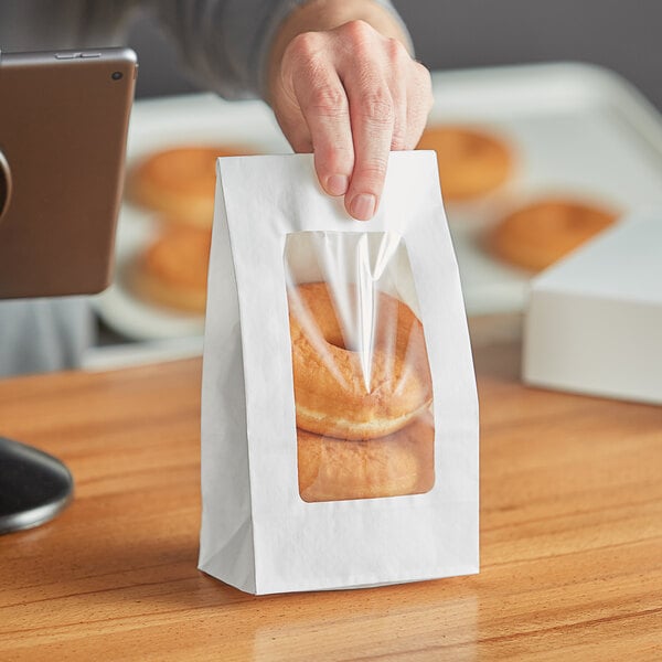 A person holding a Choice white paper bag with a clear window filled with donuts.