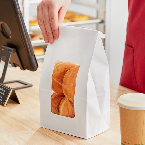 A hand holding a Choice white paper bag of doughnuts in front of a bakery display counter.