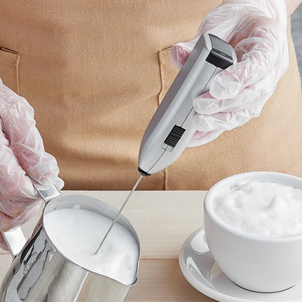 A person using a Fox Run handheld mixer to froth milk in a metal cup.