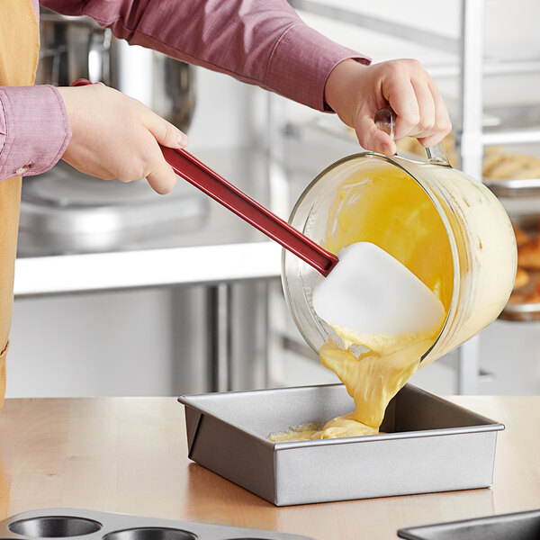 A person using a red Choice spoonula to pour yellow liquid into a pan.