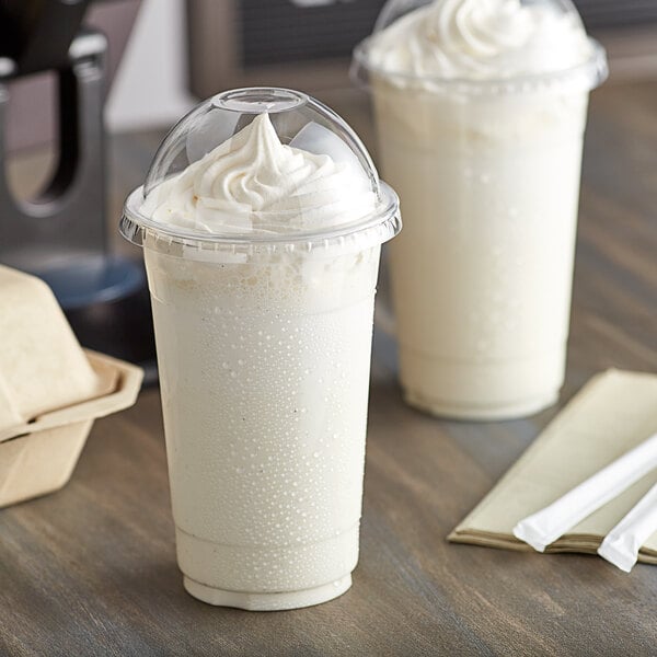 Two Choice clear plastic cups of milkshake with dome lids and straws on a table.