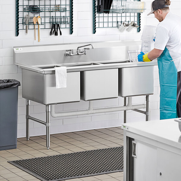 A man in a blue apron washing dishes in a commercial kitchen with a Regency 3 compartment sink.