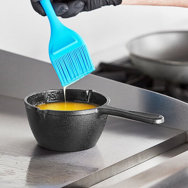 A person using a blue spatula to pour yellow liquid into a black Valor mini cast iron melting pot.