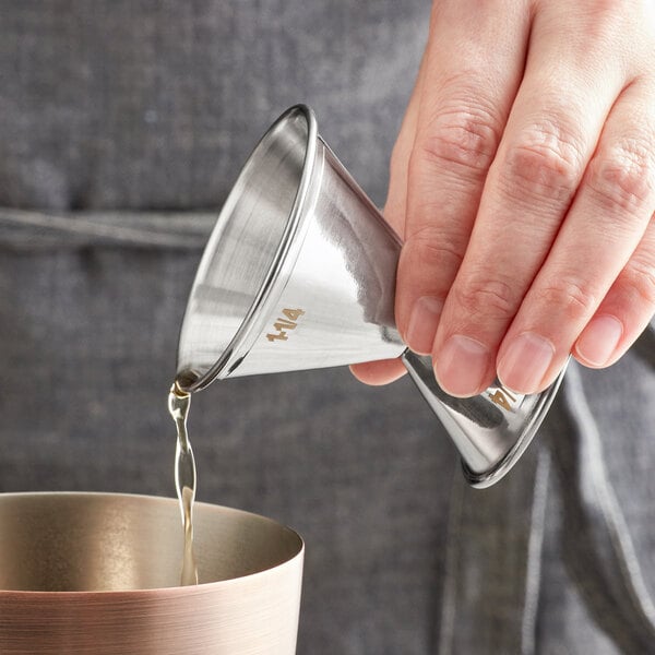 A person using an American Metalcraft stainless steel jigger to pour liquid into a cup.