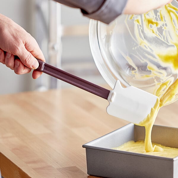 A person using a Mercer Culinary Hell's Tools silicone spatula to pour yellow batter into a pan.