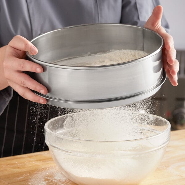 A person using a Choice stainless steel sieve to sift flour into a bowl.