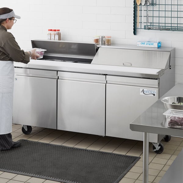 An Avantco stainless steel refrigerated sandwich prep table on a counter in a kitchen with a woman in a white apron.