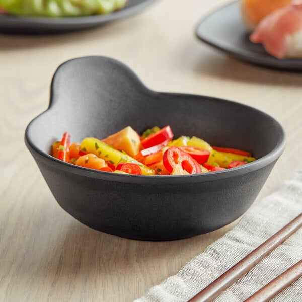 A close up of a black matte melamine bowl with handle filled with food on a table.