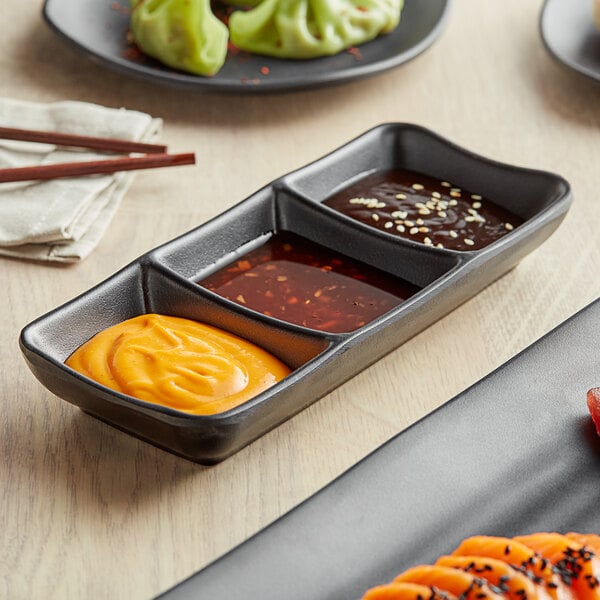 A black rectangular melamine sauce dish with three compartments filled with different sauces on a table.