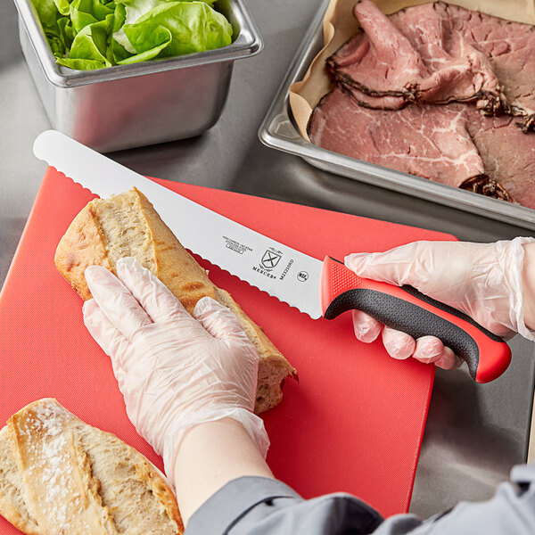 A person cutting bread with a Mercer Culinary Millennia Colors bread knife on a red cutting board.