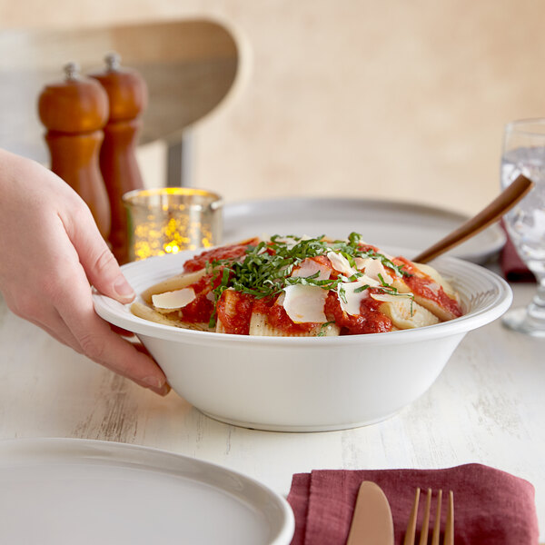 A hand holding a Tuxton white china bowl of pasta with sauce and herbs.