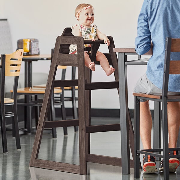 A baby sitting in a Lancaster Table & Seating bar height high chair with a wooden seat.