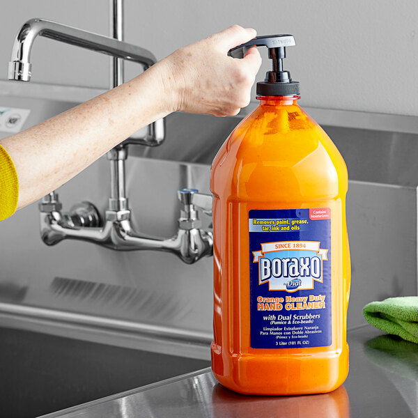 A hand holding a bottle of Dial Boraxo heavy-duty liquid hand soap with an orange label and pump.